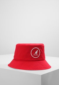 Cappello a secchiello rosso in tessuto, con un logo circolare bianco raffigurante un canguro. La visiera è larga e presenta una finitura texture.