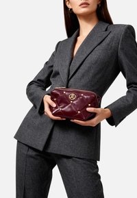 Borsa in pelle bordeaux trapuntata con dettagli dorati; presenta una chiusura con zip e borchie decorative sulla superficie; forma compatta e rettangolare.
