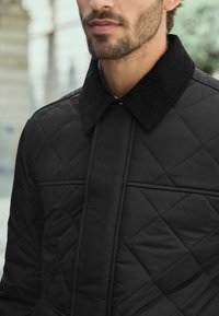 Black quilted jacket with corduroy collar, featuring diamond stitching and a front placket. Textured fabric, close-up view of neckline.