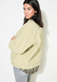 Lichtgroene oversized fleece jas met afhangende schouders, textuurstof en een geribbelde zoom, gelaagd over een gestreept shirt.