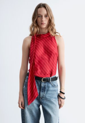 Femme aux cheveux blonds ondulés portant un haut rouge à motifs sans manches avec détail foulard, un jean bleu taille haute, une ceinture noire et des bracelets au poignet droit.