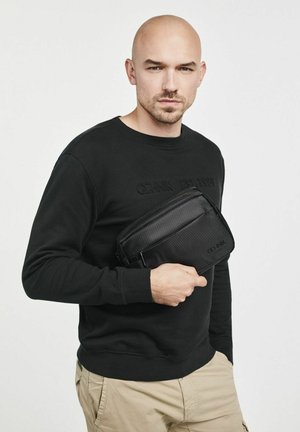 Czarna torba na ramię wykonana z teksturowanego materiału, zapinana na zamek, trzymana przy czarnej bluzie. Posiada minimalistyczny design i detal z logo.