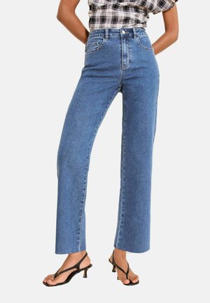 Femeie purtând blugi albaștri cu talie înaltă și croială dreaptă, asortați cu sandale negre cu barete și toc mic, și un top cu mâneci scurte în carouri.