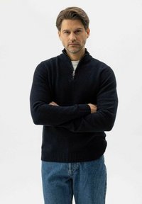 Mann mit verschränkten Armen, der einen schwarzen Reißverschluss-Pullover und blaue Jeans trägt, vor einem schlichten weißen Hintergrund.