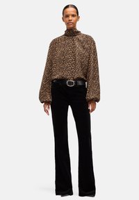 Persona che indossa una camicetta marrone a maniche lunghe con stampa leopardata e collo alto, pantaloni neri a gamba larga e cintura nera con fibbia d'argento.