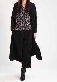 Sort lang frakke over en blomsterpintet sort bluse, parret med sorte, plisserede bredde bukser og ankelstøvler. Outfittet har et mix af mønstre og teksturer.