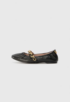 Chaussure plate en cuir noir avec des détails froncés et une bride chaîne dorée sur le dessus sur un fond gris clair.
