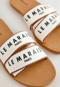 Sandales slides couleur sable avec des brides en tissu blanc ornées de coutures marron et le texte noir "LE MARAIS PARIS" sur la partie supérieure.