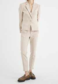 Béžový oblek na míru s přiléhavým sakem a slim kalhotami. Hladká tkanina s jemným leskem, doplněná hnědými koženými mokasíny s černými podrážkami.