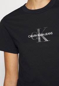 Czarny t-shirt z krótkim rękawem z błyszczącym logo "ck" i białym napisem "Calvin Klein Jeans" noszony przez osobę.