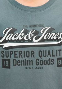 Burmārs T-kreklā izbalējušā tirkīza krāsā ar melnu drukātu tekstu: "The Authentic Jack & Jones" un "Superior Quality Denim Goods 19 90."