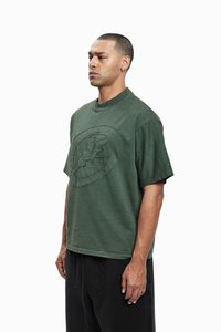 Olive green short-sleeve t-shirt with a round neckline and textured graphic design. Material appears soft, casual fit. Paired with dark pants.