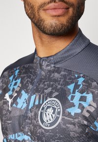 Manchester City zadrgana majica v temno sivi barvi z modrimi geometrijskimi vzorci in teksturirano tkanino; vključuje logotip in visok ovratnik.