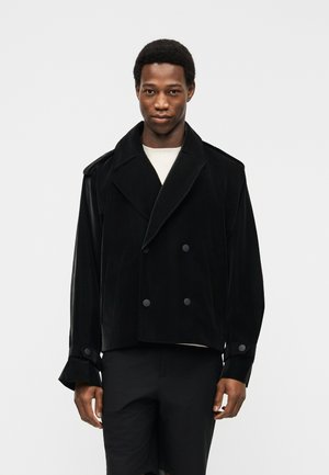 Férfi fekete dupla soros zakóban, széles hajtókával és begombolt mandzsettával, fehér inggel, egyszínű háttér előtt állva.