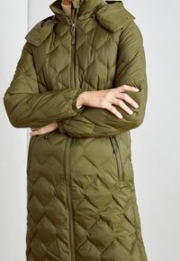 Persona vistiendo una chaqueta larga de plumas de color verde oliva con cremalleras y de pie con los brazos cruzados.