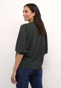 Dark green pullover with puffed short sleeves, textured fabric, and a relaxed fit. Paired with blue denim for contrast.