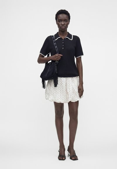 Femme portant un haut noir à manches courtes avec une finition blanche, une jupe blanche à motifs, des sandales noires et portant un sac à bandoulière noir avec des franges.