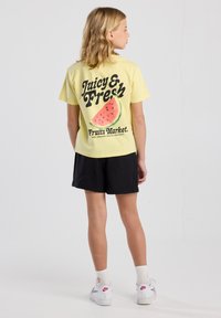 T-shirt di cotone gialla con testo "Juicy & Fresh" e grafica di anguria sul retro, abbinata a pantaloni corti neri e sneakers bianche, con un'inclinazione leggermente laterale.