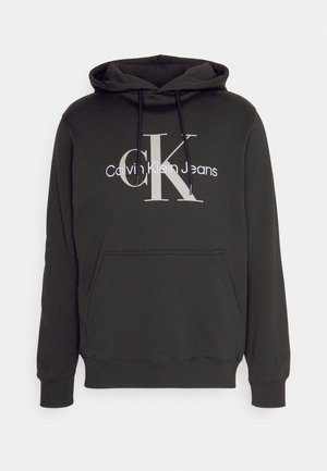 Sudadera con capucha negra hecha de tejido suave. Cuenta con un bolsillo frontal y cordones. Muestra el logo de "Calvin Klein Jeans" en blanco y gris.