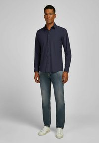Navyblauwe overhemd met lange mouwen en een reguliere pasvorm, gecombineerd met lichtblauwe jeans en witte sneakers, tegen een effen achtergrond.