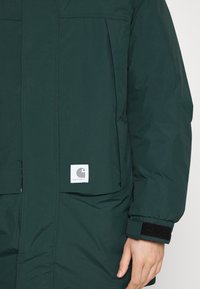 Donkergroene Carhartt-jas met zakken aan de voorkant, een zichtbaar merkembleem en een verstelbare klittenbandsluiting op de manchet, gedragen aan de rechterarm.