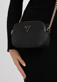 Piccola borsa a tracolla nera Guess con tracolla in catena dorata indossata sopra un vestito nero a coste, con una mano appoggiata vicino all'anca.