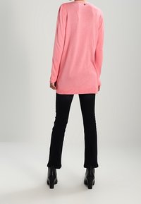 Långärmad rosa tröja med rundad halsringning, ribbade mudd, och knappparad detalj på ryggen, i kombination med svarta utsvängda byxor och ankelstövlar.