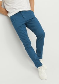 Blå bomullsbukser med smal passform, dragkedja och sidfickor, tillsammans med vita sneakers, på en ljus bakgrund.