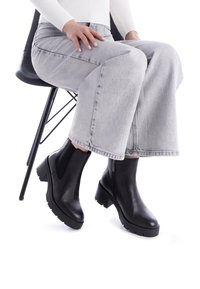 Botas de cuero negras hasta el tobillo con un tacón grueso, elástico lateral y cremallera, combinadas con unos jeans de pierna ancha gris claro.