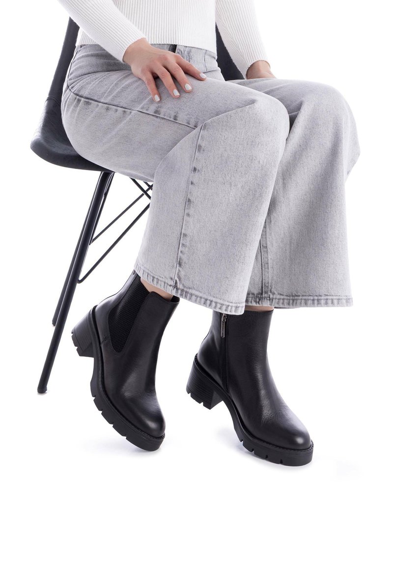 Botas de cuero negras hasta el tobillo con un tacón grueso, elástico lateral y cremallera, combinadas con unos jeans de pierna ancha gris claro.