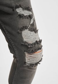 Mørke denimjeans med frynsete kanter, flere slitte partier med grått og lysere stoff under, som viser en uregelmessig tekstur.