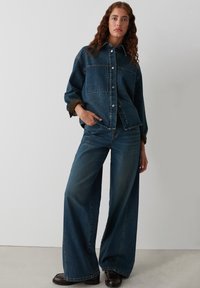 Denim jas en wijde jeans in donkerblauw, met een ontspannen pasvorm, grote zakken en zilveren knopen. Glad textuur met gestikte details.