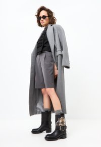 Abrigo largo gris sobre una blusa negra, pantalones cortos grises y botas de tobillo negras con tachuelas en forma de pirámide. Los zapatos tienen un diseño estructurado y un tacón plano.