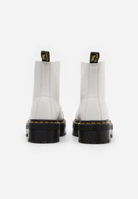 Botas de tobillo de cuero blanco con un acabado texturizado, suelas de plataforma negras y costuras amarillas. Cuentan con una lengüeta en la parte posterior para ponérselas fácilmente.