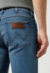 Jeans in denim blu con un'etichetta in pelle marrone sul taschino posteriore e cuciture a contrasto. Dettaglio ravvicinato di un design aderente e di un tessuto testurizzato.