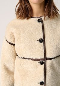 Giacca in pelliccia sintetica beige con una forma arrotondata, accenti marrone scuro, cinque bottoni neri e dettagli cuciti lungo i bordi.