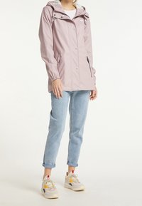 Veste imperméable rose avec capuche, poches zippées, boutons pressions et poignets élastiques. Associée à un jean bleu clair retroussé et des baskets de sport.