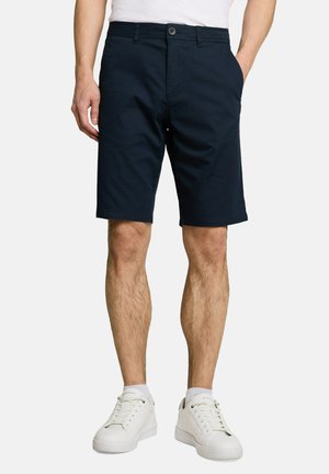 Hombre con pantalones cortos azul marino, camisa blanca metida dentro, calcetines blancos y zapatillas blancas de pie sobre un fondo neutro.
