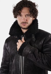 Giubbotto in shearling nero con un colletto e polsini morbidi e testurizzati. Presenta una zip centrale e clip regolabili sul colletto.