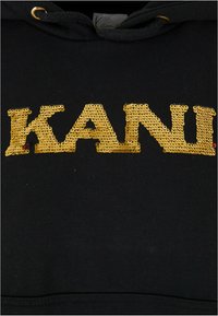Čierna mikina s veľkým zlatým flitrovým nápisom "KANI." Materiál sa zdá byť mäkký, s kapucňou na šnúrku a bežným strihom.