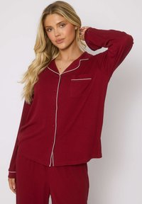 Completo Pigiama bordeaux realizzato in tessuto a costine, composto da una maglia con bottoni, taschino sul petto e piping bianco a contrasto, abbinato a pantaloni coordinati.