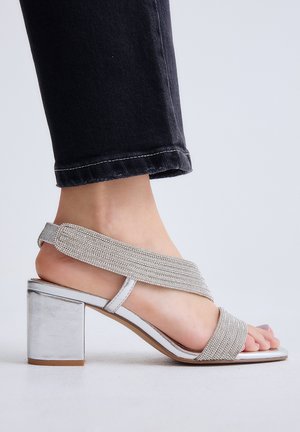 Pie con sandalia plateada de tacón cuadrado con tiras de piedras brillantes combinada con vaqueros negros arremangados, sobre una superficie blanca.