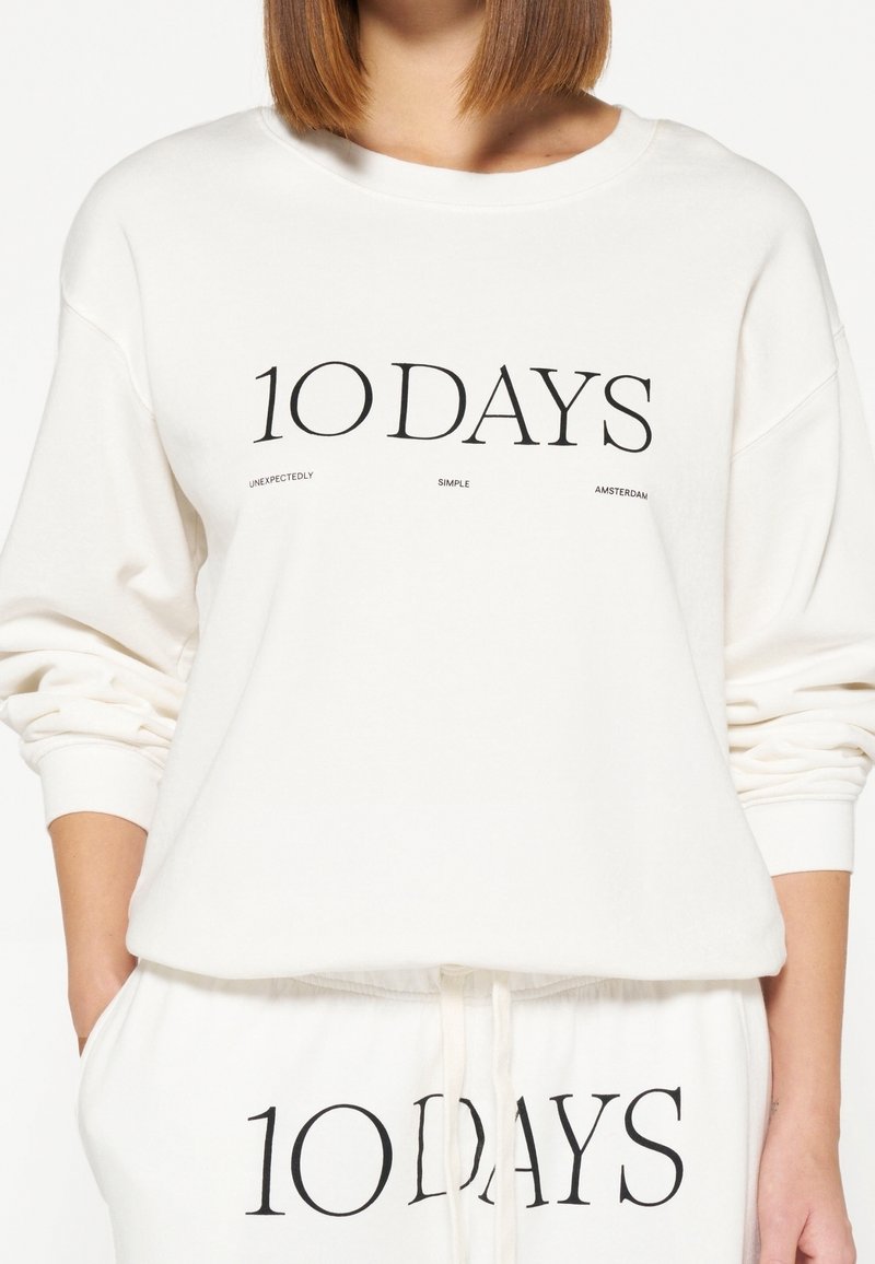 Sudadera blanca con texto negro "10 DAYS" en el frente. Presenta hombros caídos, ajuste relajado y puños acanalados. Combínala con pantalones a juego.