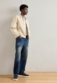 Beige lett jakke med elastiske mansjetter, hvit skjorte, blå slitte jeans og blå slip-on sko med hvit såle, stående på tregulv.