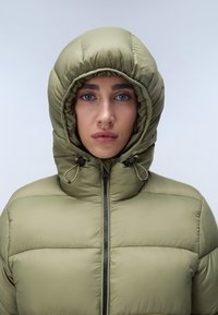Olijfgroene pufferjack met een aansluitende capuchon. Voorzien van een ritssluiting en aantrekkoorden. Textuurstof met gequiltte secties.