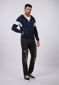 Veste à zip marine avec des rayures blanches, en mélange de coton, poches frontales, assortie à un jean noir dégradé et des baskets noires.