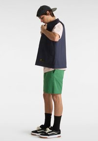 Mørkeblå ærmeløs vest med lommer, iført over en lys pink skjorte; parret med grønne shorts og sorte sneakers med hvide såler.