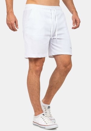 Man draagt witte shorts met trekkoord en witte lage sneakers, staat met de benen iets uit elkaar tegen een effen achtergrond.