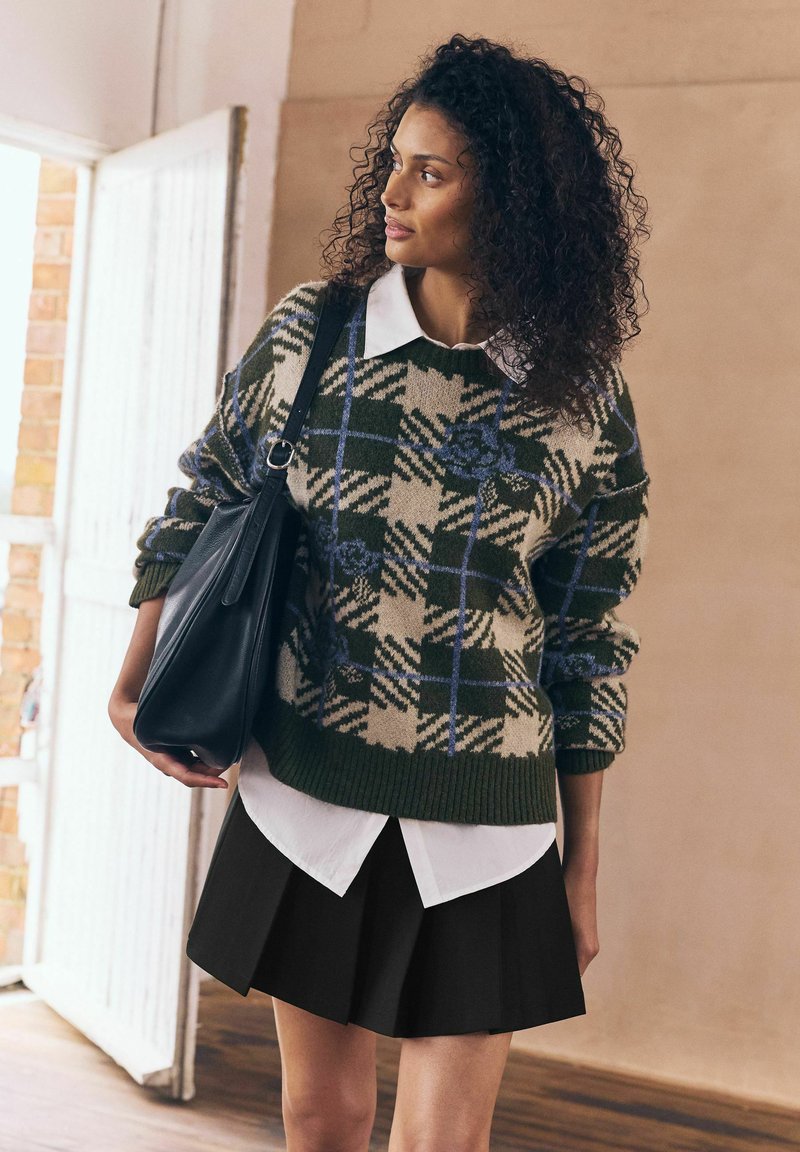Woman with curly hair wearing green patterned sweater, white collared shirt, black skirt, and carrying a black leather shoulder bag indoors.