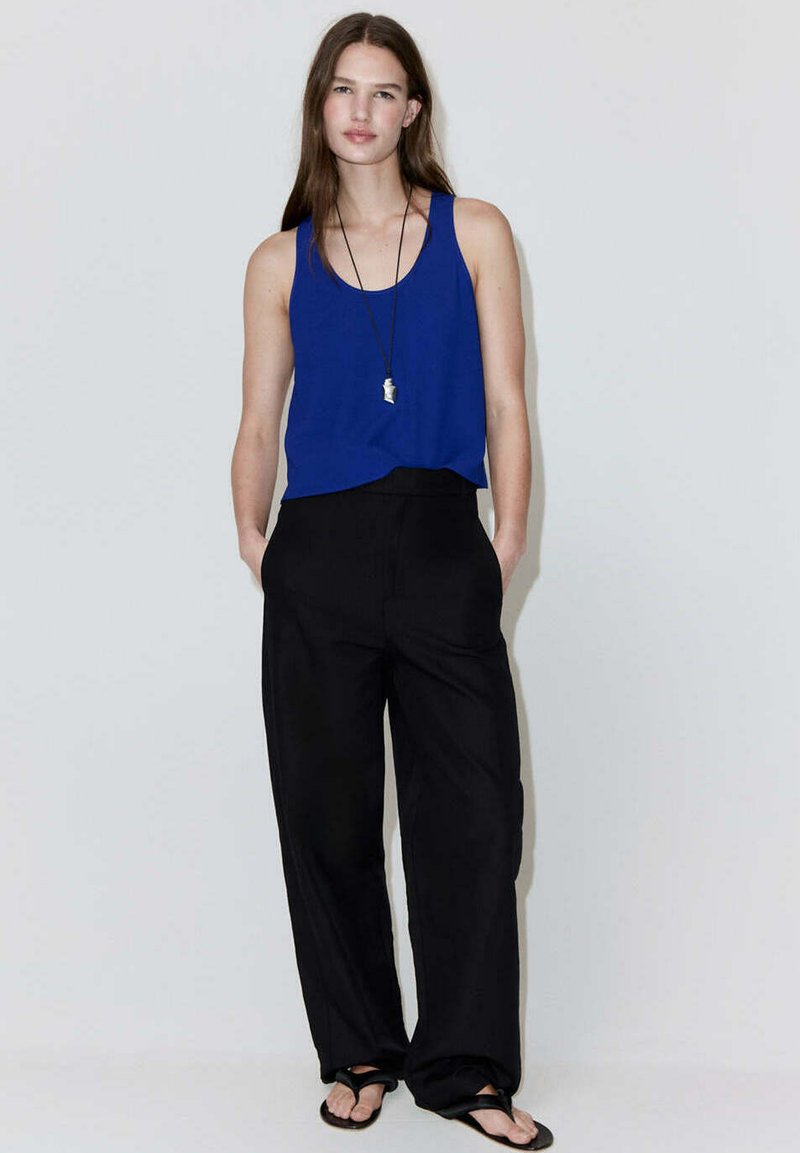 Jeune femme aux longs cheveux portant un haut bleu sans manches, un pantalon noir à jambes larges, des sandales noires et un collier pendentif, debout, détendue.
